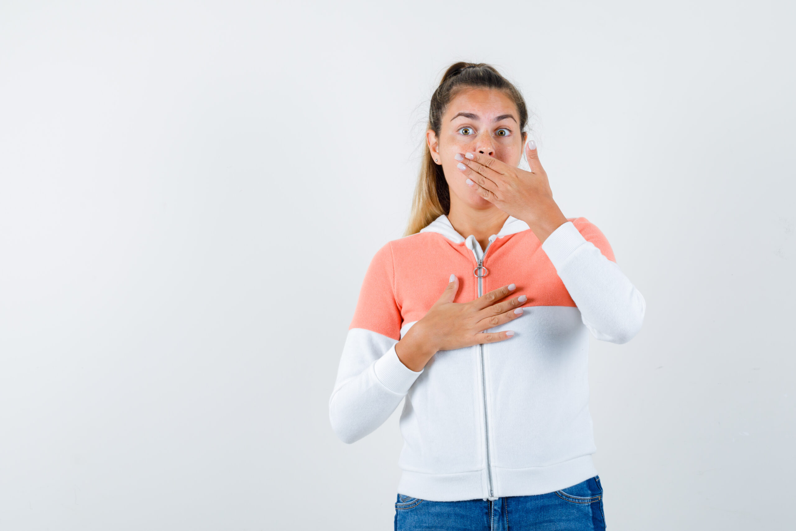 Portrait of young lady keeping hand on mouth in jacket and looking scared front view